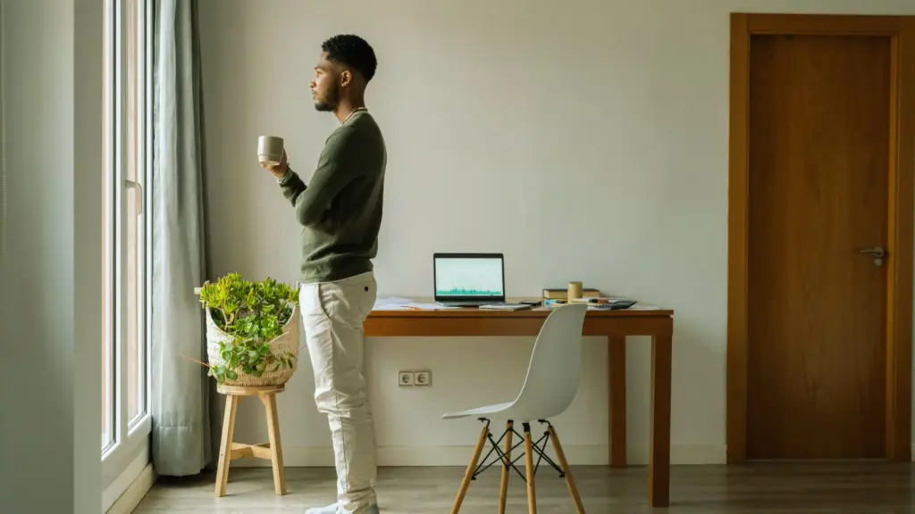 A man standing indoors, holding a cup of tea, thoughtfully gazing out the window