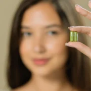A young woman holding two green supplement capsules between her fingers, looking at them with a thoughtful expression A young woman holding two green supplement capsules between her fingers, looking at them with a thoughtful expression